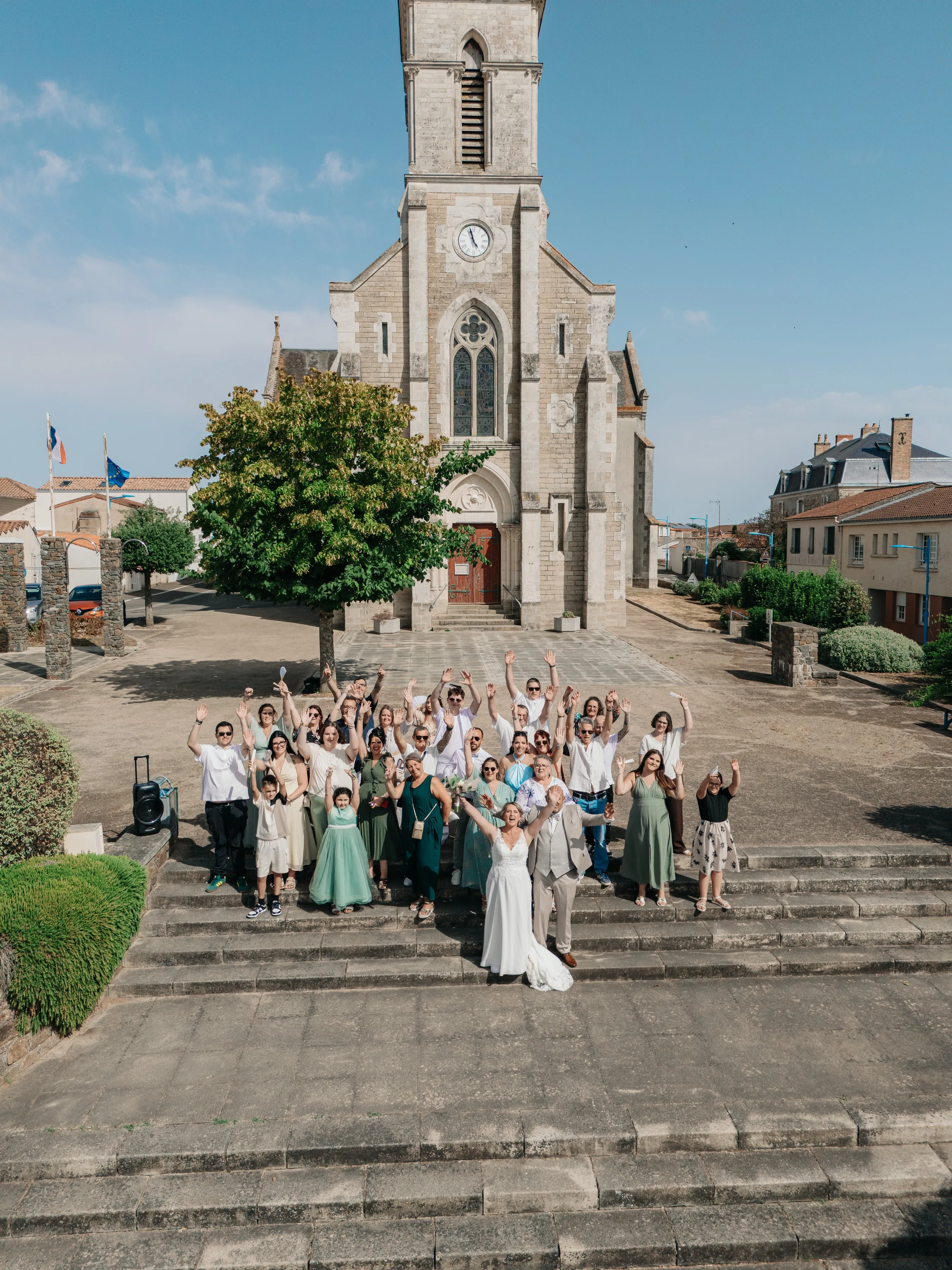Les mariés et leurs invités, bras levés sur les marches de l’église de Vairé, vus du ciel