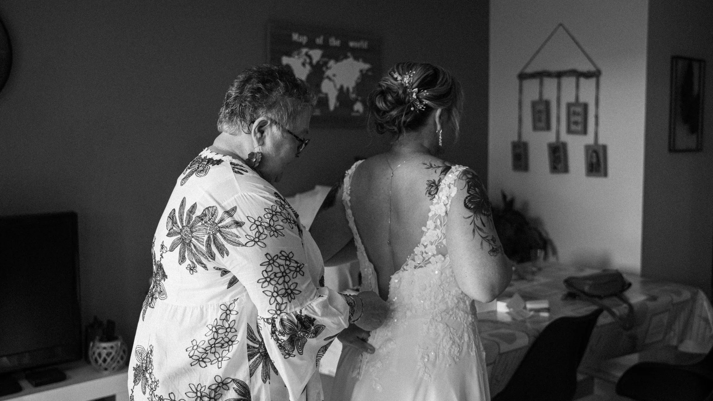 Une proche aide la mariée à fermer sa robe, en noir et blanc