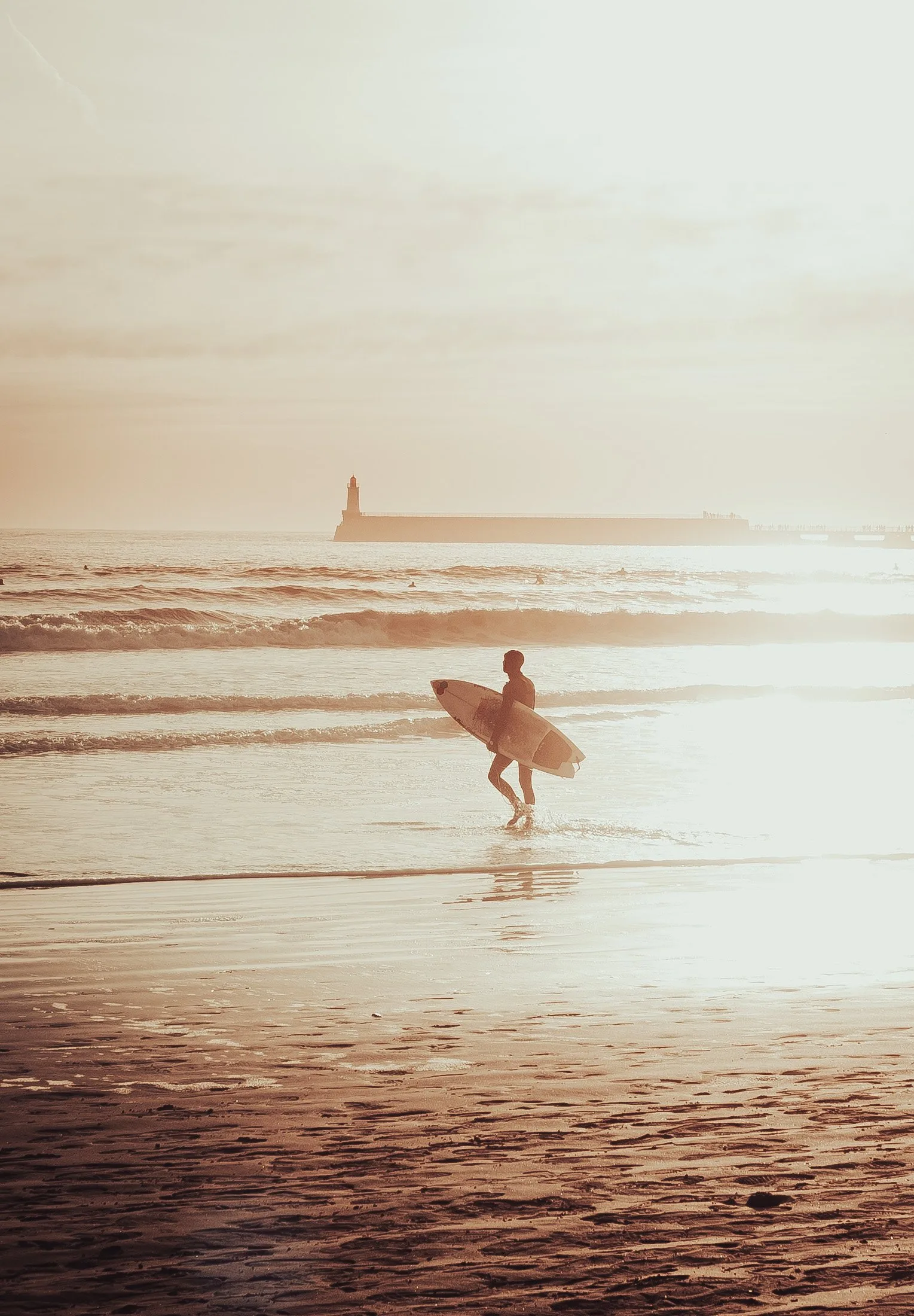 Un surfeur sort de l’eau au soleil couchant, le phare des Sables-d’Olonne au loin