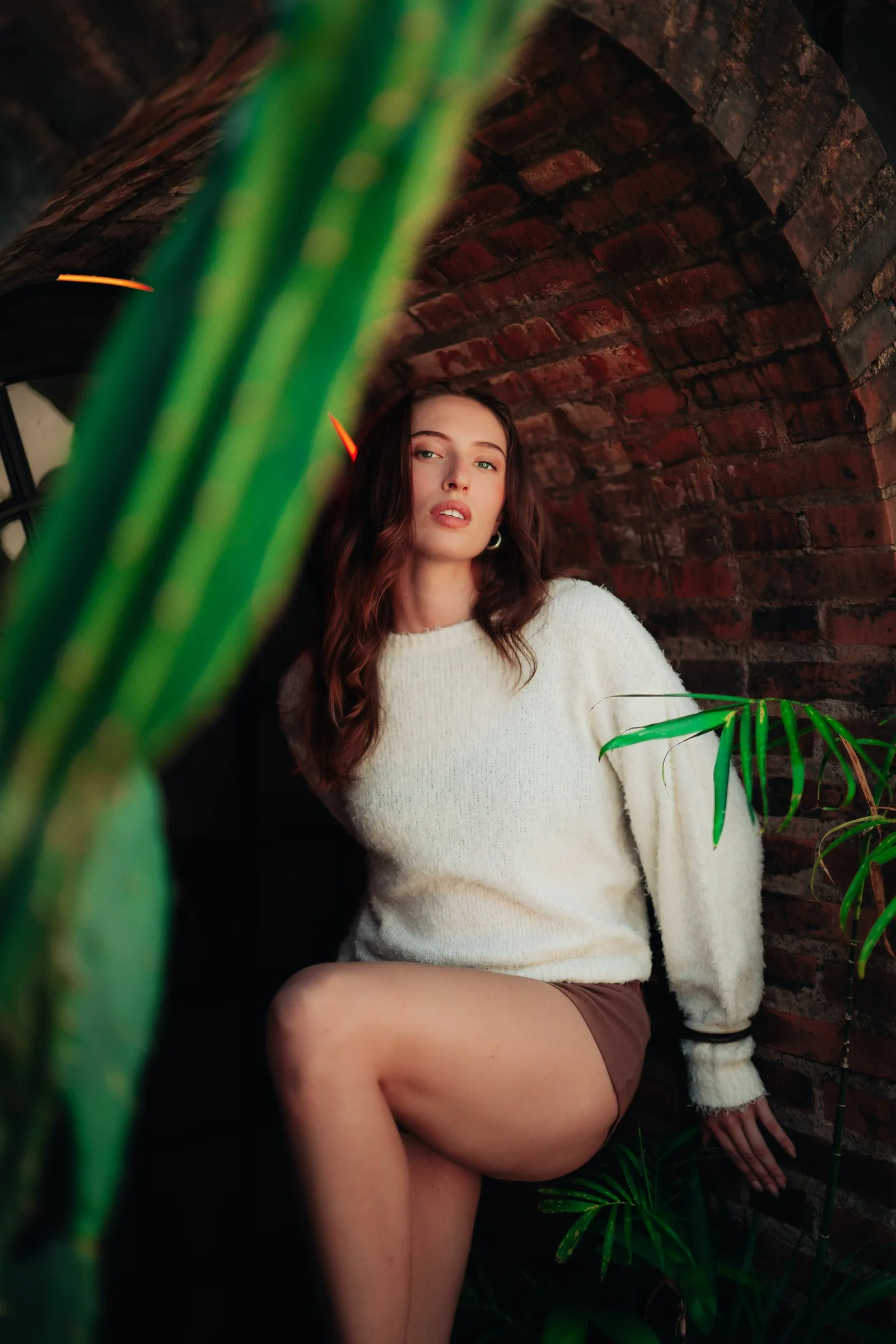 Femme en pull blanc parmi les plantes, devant un mur de briques