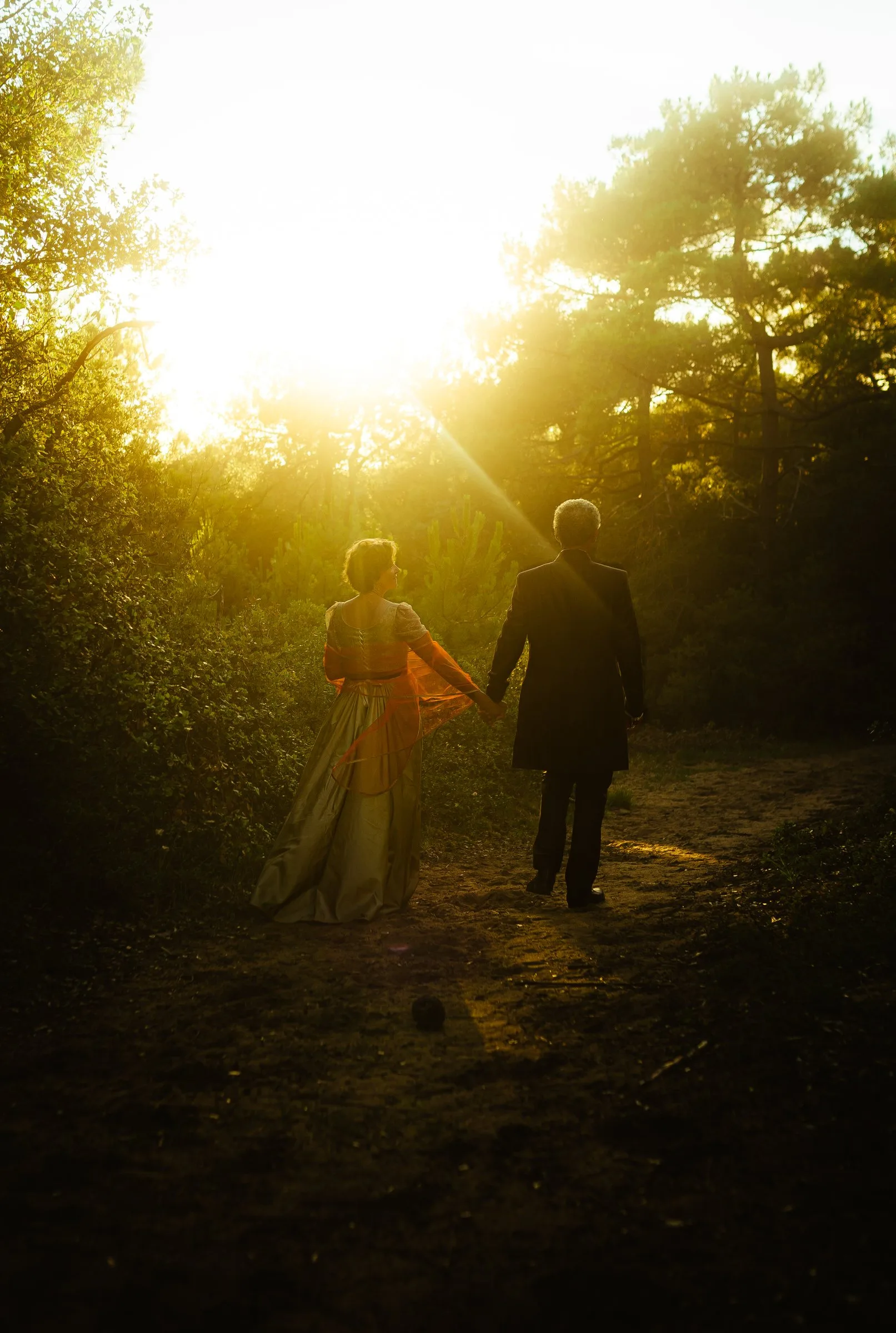 Pascale et Louis marchent main dans la main sur un sentier sous les pins, à contre-jour